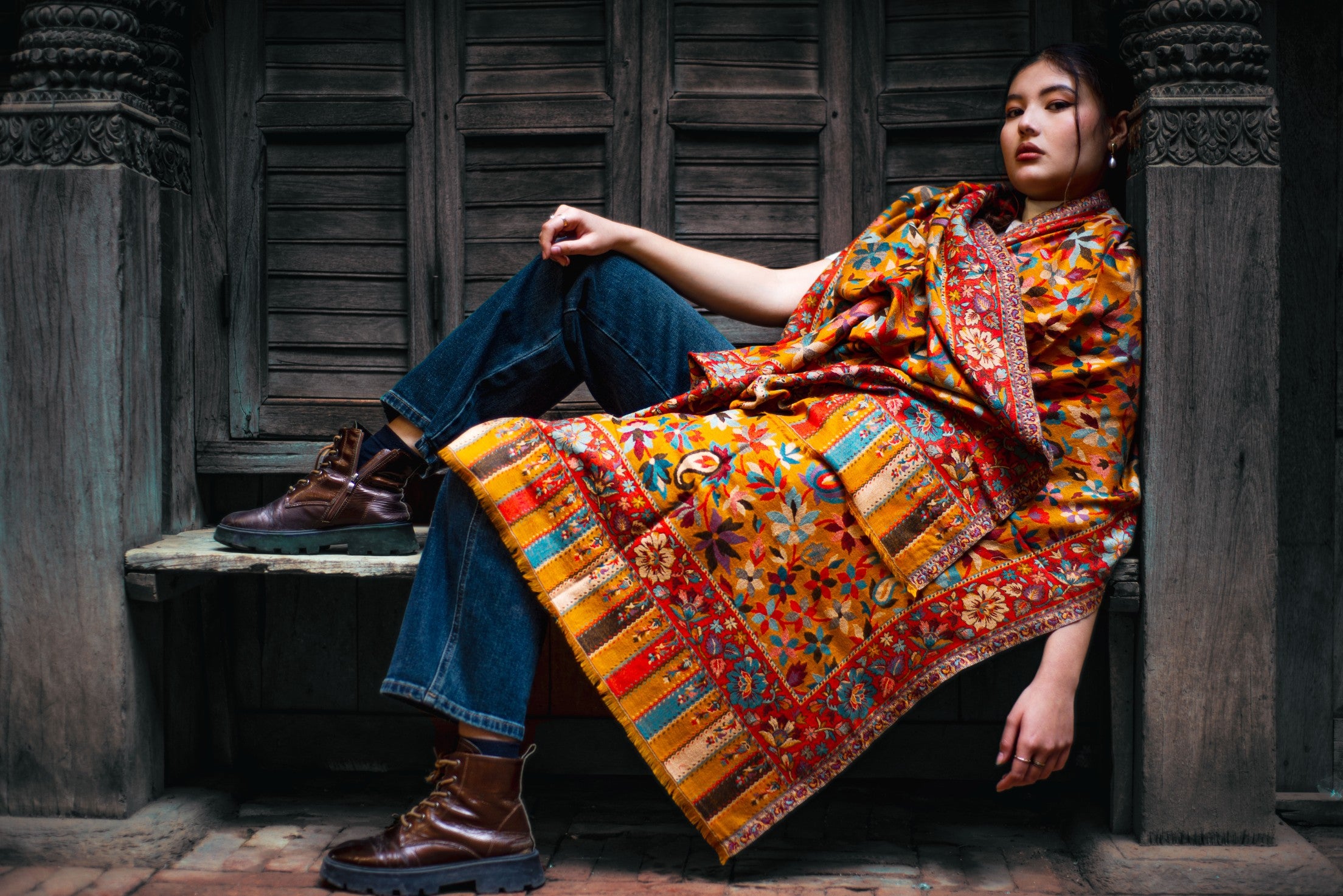Person wearing a colorful patterned shawl sitting against a wooden wall.